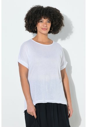 Kvinde med krøllet hår iført store gyldne øreringe, en løs hvid kortærmet top og et sort nederdel, der står foran en hvid væg.