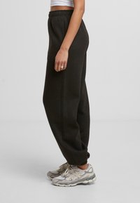 Personne portant un pantalon de survêtement sombre, un haut court blanc, des baskets beiges et grises, se tenant de côté contre un fond clair uni.