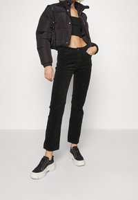 Veste matelassée noire, coupe courte, portée par-dessus un haut noir ajusté. Pantalon en velours côtelé noir et baskets noires épaisses avec semelles blanches.