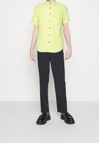 Krótki rękaw, jasn żółta koszula z guzikami o teksturowanej tkaninie, w połączeniu z ciemnymi, dopasowanymi spodniami i masywnymi czarnymi butami.