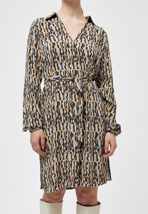 Kobieta w beżowej sukience do kolan z abstrakcyjnymi czarnymi i niebieskimi wzorami, z kołnierzykiem, długimi rękawami i paskiem wiązanym w talii.