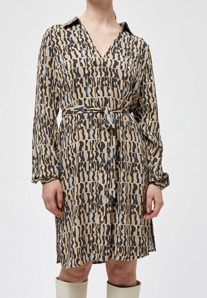 Femme portant une robe beige longueur genou avec des motifs abstraits noirs et bleus, un col, des manches longues, et une ceinture nouée à la taille.
