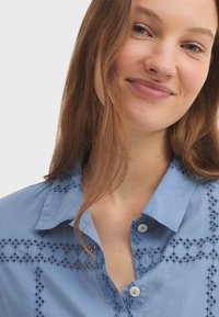 Chemise en coton bleu clair avec col boutonné, détails en dentelle découpée au laser et boutons contrastants. Texture lisse, design décontracté.