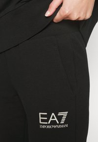Main avec un vernis à ongles clair tire sur une ceinture noire ; le logo EA7 Emporio Armani apparaît sur un pantalon noir près de la poche.