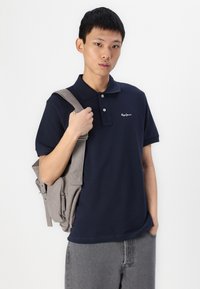 Jūras spēku polo krekls ar īsām piedurknēm, divu pogu apkakle, izšūts logo. Apvienots ar pelēku auduma somu ar rāvējslēdzējiem un regulējamu siksnu.