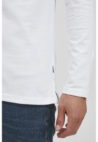 Hvit langermet skjorte med rund hals, laget av glatt bomull, har sideslitser i nederkant, brukt over mørke denimjeans.