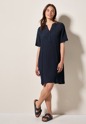 Robe bleu marine à manches courtes avec un décolleté en V, présentant un motif texturé et froissé. Associée à des sandales à glissière foncées reposant sur une surface claire.