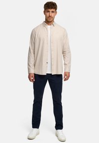 Hellbeige Hemd mit Knöpfen, getragen über einem weißen T-Shirt, kombiniert mit dunkelblauen Hosen und weißen Turnschuhen. Das Hemd hat eine lockere Passform.