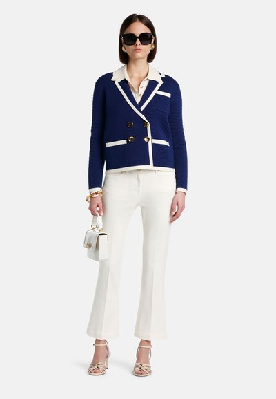 Sieviete stāv, valkājot tumši zilu dubultpogāju žaketi ar baltu apmali, baltas bikses, bēšas augstpapēžu kurpes, lielas melnas saulesbrilles un turot baltu somiņu.