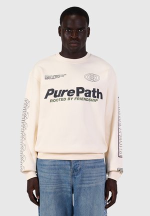 Man draagt een crème sweatshirt met de tekst "Pure Path Rooted by Friendship", met patroon op de mouwen en een blauwe spijkerbroek tegen een effen lichte achtergrond.