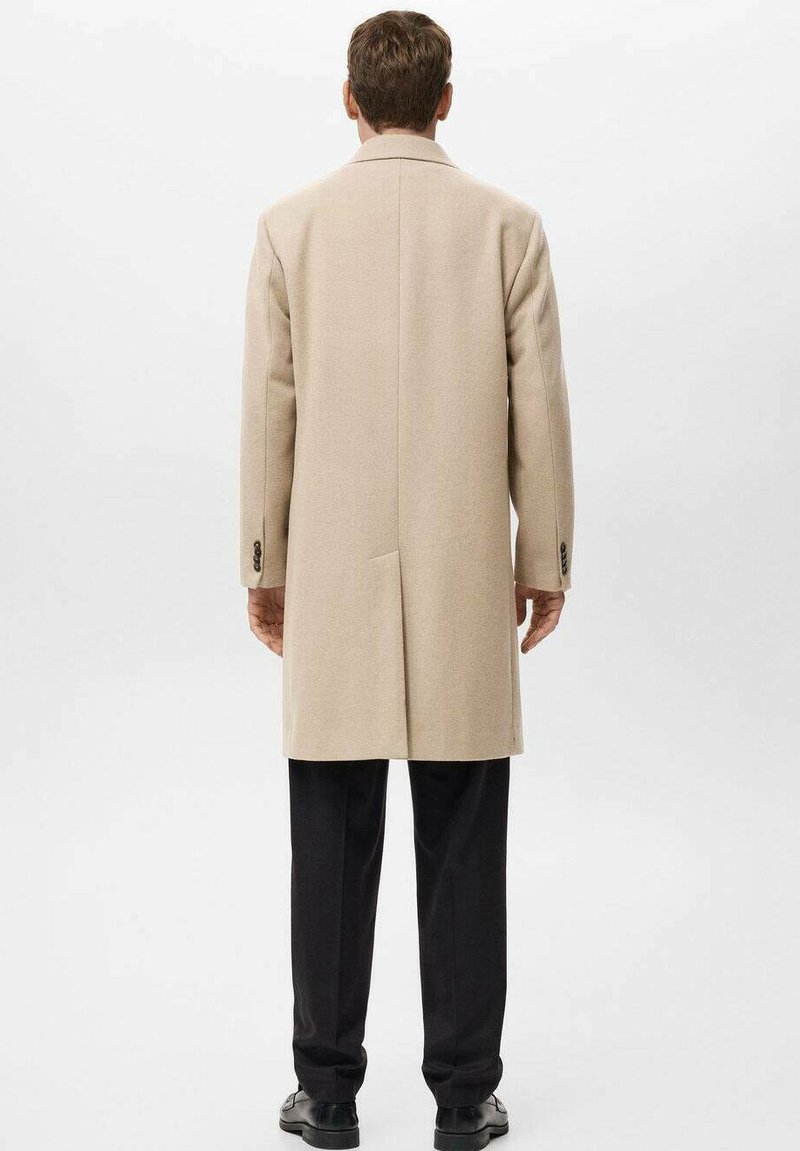 Beige wollen jas met een rechte snit, gelletjes kraag en lange mouwen; heeft een enkele split aan de achterkant en zwarte broeken.