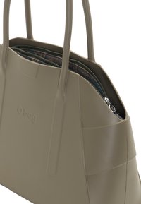 Tummanvihreä kantokassi, jossa on kaksinkertaiset kahvat, sileä kumimainen pinta, vetoketjukiinnitys ja kuviollinen sisus. Etupuolella on kohokuvioitu logo.