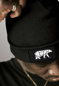 Man adjusting black knit beanie with geometric white bear logo patch above his eyebrow, wearing black shirt and gold chain.