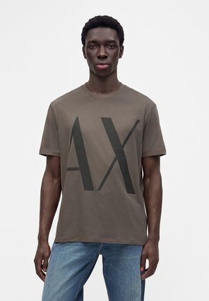 Giovane uomo dalla pelle scura che indossa una maglietta marrone con grandi lettere nere "AX" e jeans blu, in piedi davanti a uno sfondo chiaro e uniforme.