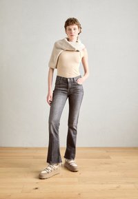 Débardeur beige, jeans évasés gris et baskets compensées beiges. Pull en tricot posé sur une épaule. Sol en bois et mur neutre en arrière-plan.