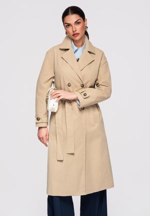 Frau trägt beigen Trenchcoat über hellblauem Hemd und dunkler Hose, hält kleine weiße Handtasche vor hellgrauem Hintergrund.