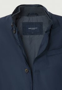 Marinblå jacka med ståkrage, knäppning fram, inre grå herringbone-foder och Hackett London-märke i storlek medium.