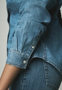 Jeanshemd aus hellblauem Stoff, mit knöpfbaren Manschetten und lockerer Passform. Strukturierte Oberfläche mit subtilen Auswaschungen und Kontrastnähten.