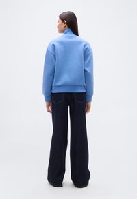 Ανοιχτό μπλε φούτερ με ψηλό γιακά, ραμμένα στους ώμους, και ριμπ μανσέτες, συνδυασμένο με σκούρο τζιν φαρδιές παντελόνες, από πίσω όψη.