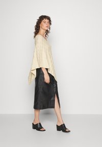 Poncho bege tricotado com um padrão de malha aberto, combinado com uma saia de couro preta até aos joelhos com uma fenda lateral, e mules pretos de salto.