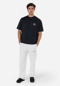 Black cotton t-shirt with circular logo on the chest, paired with white sweatpants featuring embroidered text, and black sneakers.