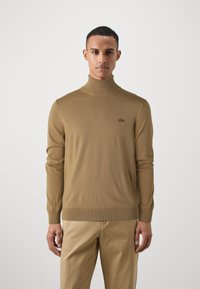 Tan spalvota turtleneck megztinis, pagamintas iš lygaus megztinio audinio, su prigludusia gamyba, raštuotais rankogaliais ir apatine dalimi, su mažu logotipo ženklu ant krūtinės.