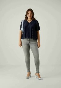 Mornarsko modra plisirana majica s kratkimi rokavi; svetlo sivi usnjeni hlače; sijoče srebrne pete; minimalistična svetla ozadja.