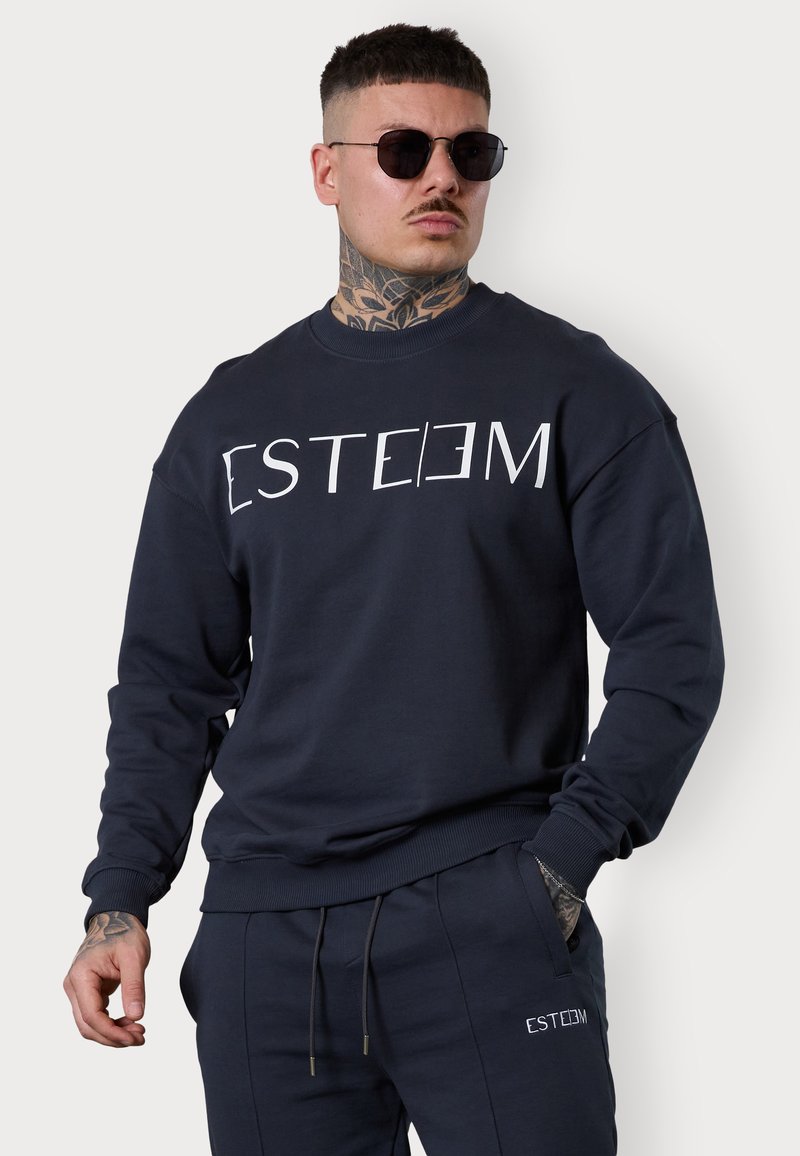 Sweatshirt bleu marine foncé avec le texte blanc "[ESTEEM]" sur le devant, présentant un col rond et des poignets côtes, accompagné d'un bas de jogging foncé assorti.