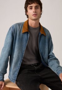 Νέος άνδρας καθισμένος σε ξύλο παγκάκι, φοράει μπλε τζιν μπουφάν με καφέ γιακά, σκούρο γκρι πουκάμισο και μαύρο παντελόνι, με απλό φόντο.