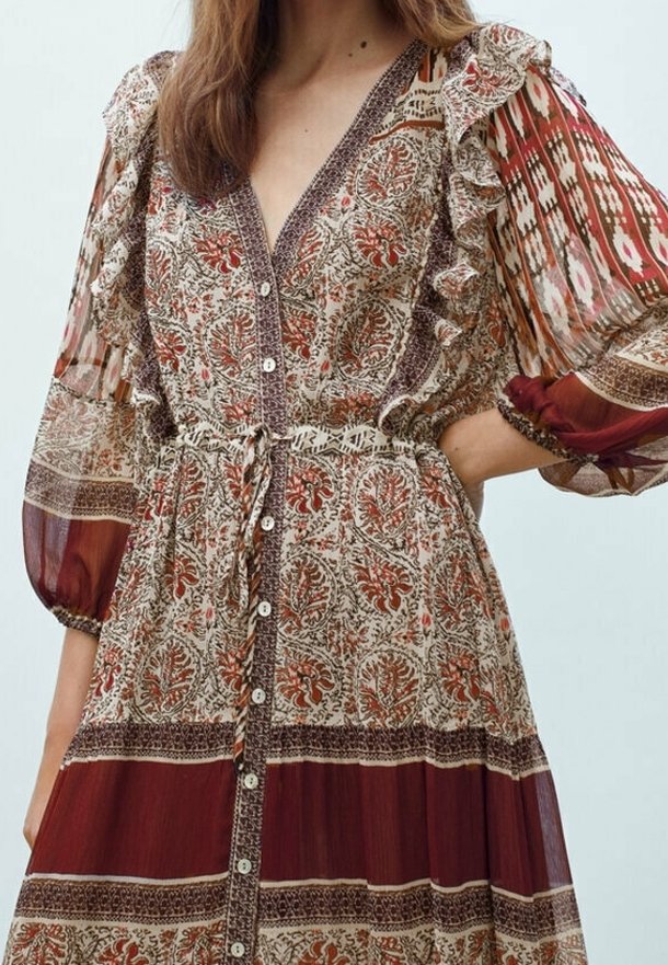 Femme portant une robe à motifs avec des épaules volantées, une patte de boutonnage à l'avant et une taille cintrée présentant des motifs floraux et géométriques dans des tons rouges et bruns.