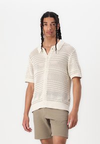 Polo shirt a maniche corte lavorato a maglia in crema con design a trama aperta. Presenta un colletto piatto, chiusura con bottoni e polsini a costine; abbinato a pantaloni corti beige.