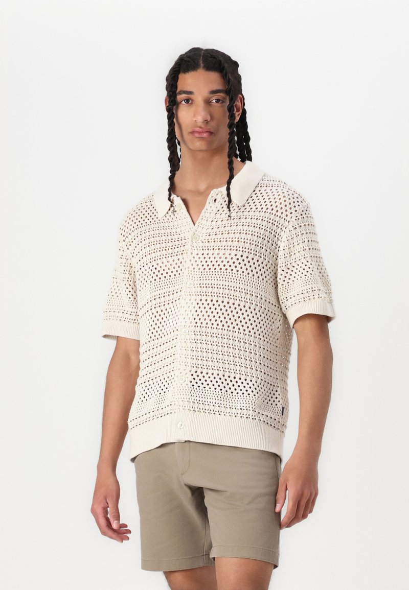 Polo shirt a maniche corte lavorato a maglia in crema con design a trama aperta. Presenta un colletto piatto, chiusura con bottoni e polsini a costine; abbinato a pantaloni corti beige.