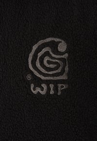 Tessuto di pile nero con un prominente logo ricamato grigio. La texture è morbida con un aspetto leggermente peloso.