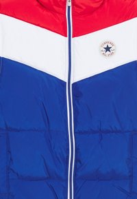 Chaqueta de plumas azul con diseño de chevrones rojos y blancos. Cierre de cremallera con acentos blancos y un parche redondo del logo de Converse en el pecho.
