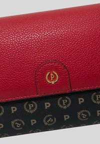 Chiusura a pattina in pelle rossa testurizzata con dettaglio del logo dorato; sezione inferiore nera con motivo a emblema dorato ripetuto.