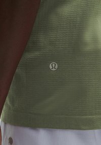 Olive green athletic top with a textured finish, featuring a small white logo on the lower left side. Short, half-length design.