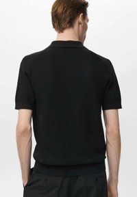 Polo shirt nero a maniche corte, in tessuto a coste testurizzato, con colletto classico e orlo aderente. Presenta estremità delle maniche arrotolate e dettagli sottili.