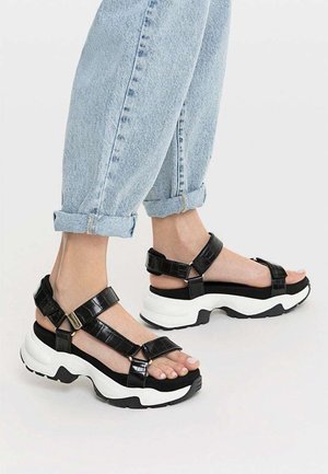 Pieds portant des sandales à plateforme noires avec des semelles blanches épaisses et des lanières réglables, associées à un jean bleu clair retroussé.