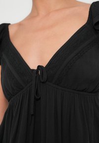 Μαύρο φόρεμα με V-λαιμόκοψη, υφή υφάσματος, λεπτομέρειες δαντέλας, πιέτες και δέσιμο στο μπροστινό μέρος. Απαλή, ρέουσα σιλουέτα.