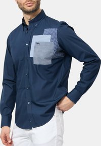 Camicia a maniche lunghe blu navy con colletto a bottoni, dotata di tre tasche a toppa in tessuto a pois blu chiaro e blu navy.