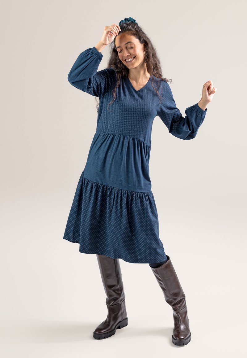 Blaues gestricktes Kleid mit einem gestuften Design, langer Ärmel, V-Ausschnitt und strukturiertem Oberteil. Getragen mit braunen Kniehohen Stiefeln.