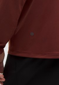 Röd långärmad idrottsskjorta med en mjuk yta, rundad nederkant och en liten logotyp i nedre vänstra hörnet. Kontrasterar med svarta byxor.