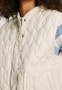 Prošívaná krémová vesta se zapínáním na patentky, s texturovaným diamantovým vzorem a přední kapsou. Pod ní je modro-bílá pruhovaná košile barevně sladěná.