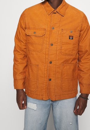 Mand iført orange manchesterjakke med fire forlommer og knaplukning, kombineret med lyseblå revnede jeans.