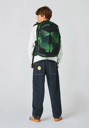 Junge im Profil mit schwarzem und grünem geometrischem Rucksack, weißem Hemd, dunkler Hose mit lächelndem Aufnäher und braunen Schuhen.