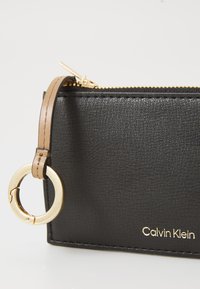 Černá texturová kožená peněženka se zlatým zipem, zlatým kroužkovým akcentem a béžovým popruhem. Obsahuje embosované logo "Calvin Klein" ve zlaté barvě.