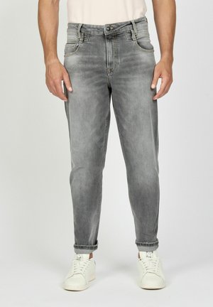 Mann trägt ausgebleichte graue Jeans, an den Knöcheln hochgekrempelt, und weiße Sneakers, steht vor einem schlichten Hintergrund.