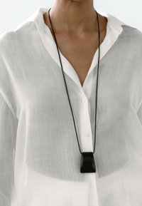 Collana a pendente nera con design geometrico, sospesa su un cordino nero, indossata su una camicia bianca trasparente con bottoni.