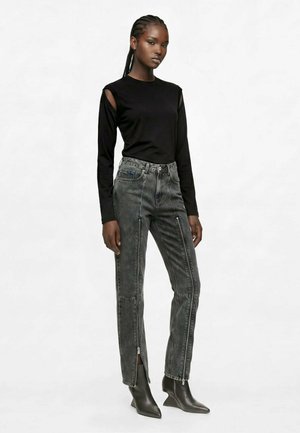 Femme aux cheveux tressés portant un haut noir à manches longues avec des découpes aux épaules, un jean gris taille haute avec des fermetures éclairs à l'avant, et des bottes noires à bouts pointus.