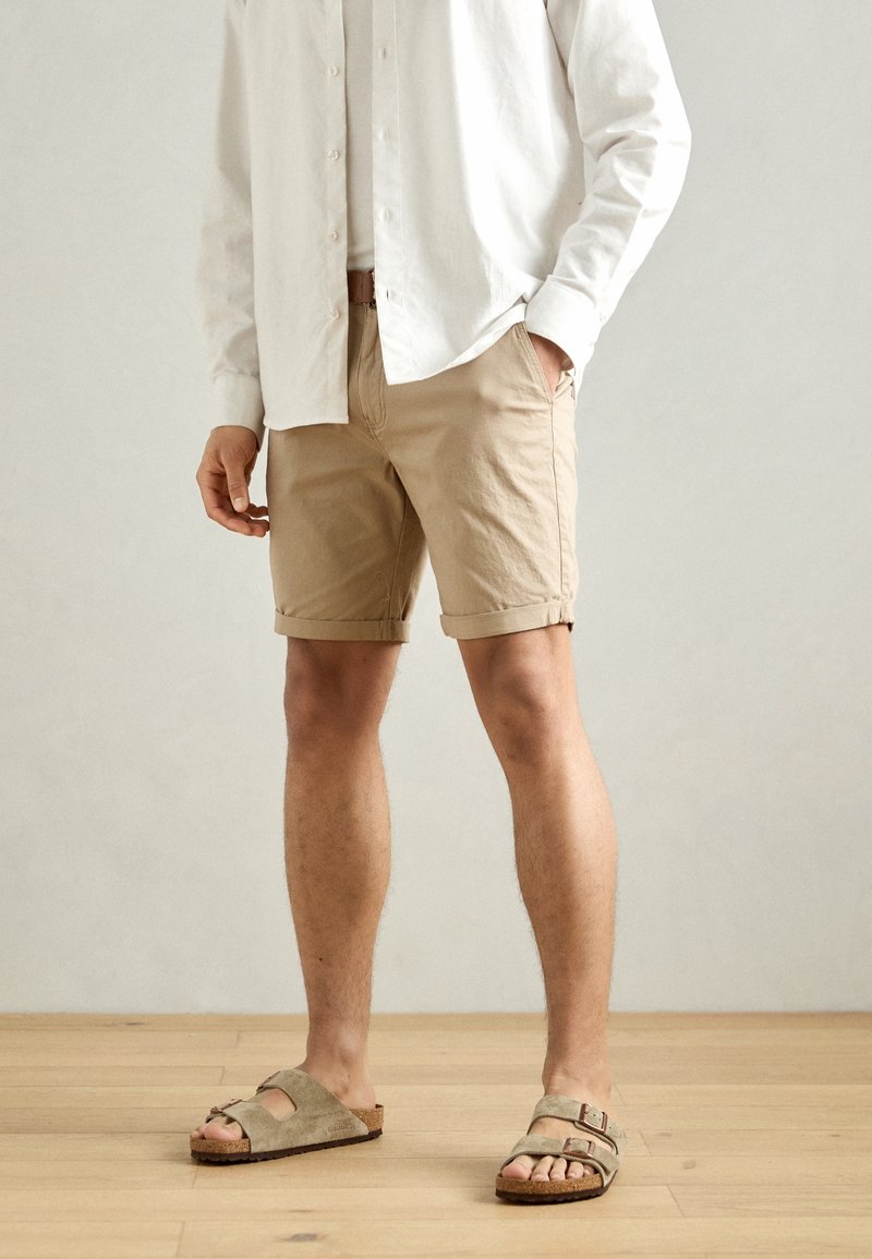 Shorts in cotone beige con risvolti, tasche frontali, abbinati a sandali in suede grigi con cinturino a fibbia, indossati con una camicia bianca a bottoni.